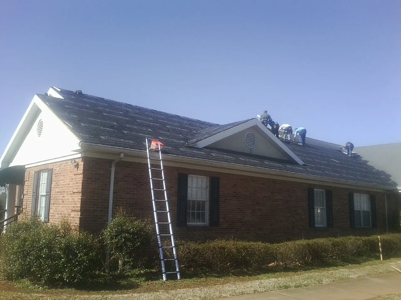 Crew tearing shingles off a brick ranch roof with a ladder set — Metal Roof Repair work by our crew in Elberta