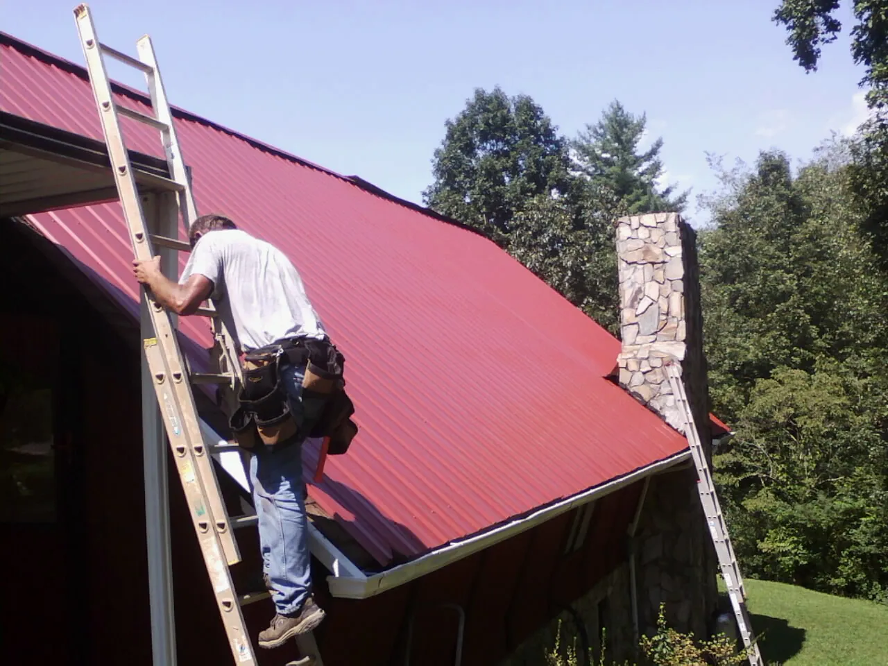 Roofer on a ladder beside a red metal roof and stone chimney — Soffit & Fascia Repair by Roofing Brothers in Elberta